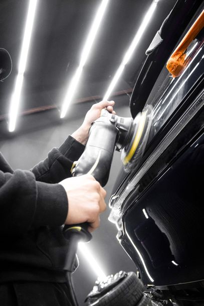 Person polishing a black car with a buffer under bright garage lights