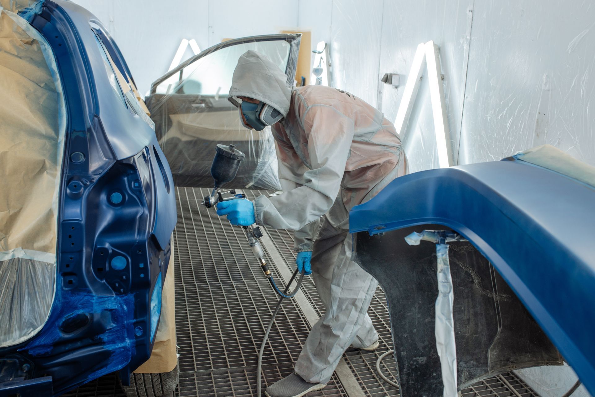 Worker in protective gear spray-painting a blue car body in a paint booth