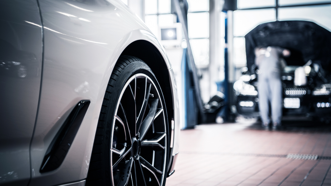 Silver car's wheel in a car repair shop. A mechanic works on another car in the background.
