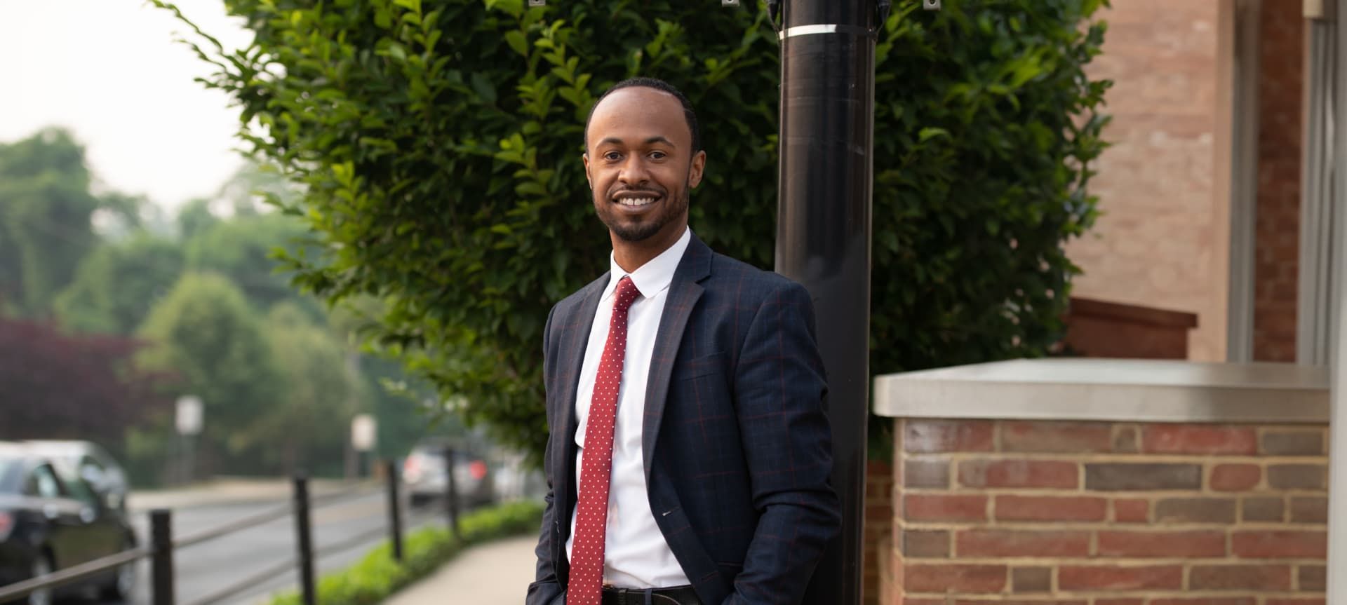 A man in a navy suit and red tie smiling while standing outdoors next to a brick wall and green foliage.