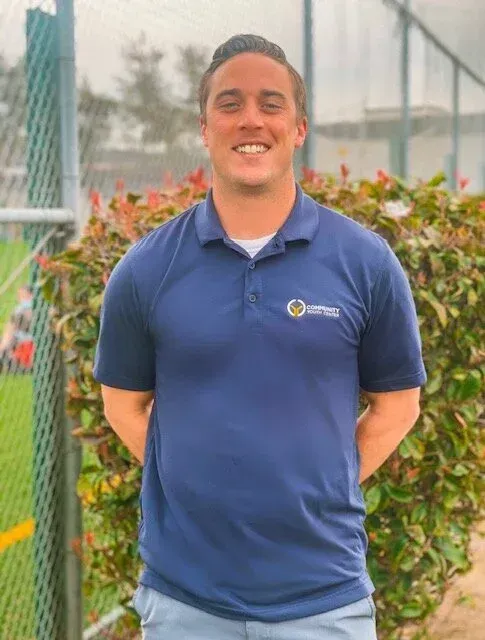 Man in blue polo shirt smiles, stands in front of green bushes and a chain link fence.