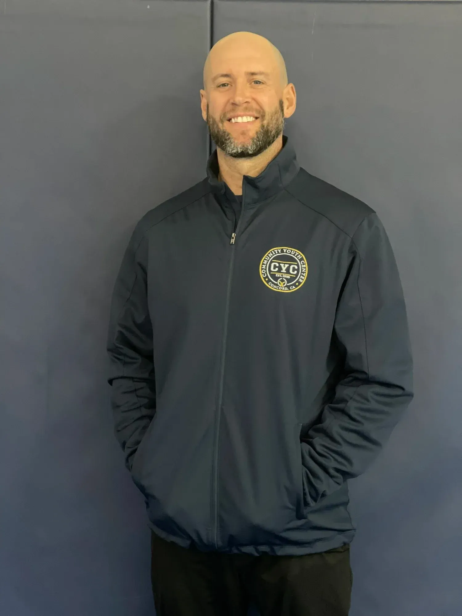 Man in navy blue jacket smiling, standing in front of a blue wall.