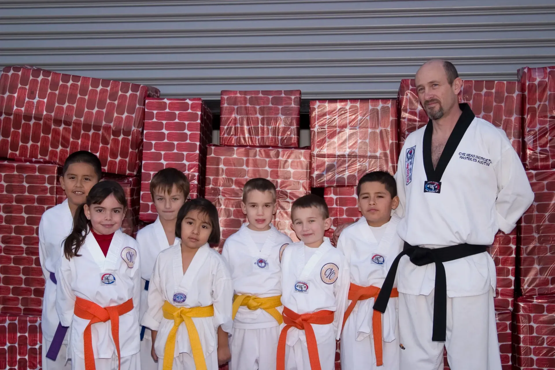 Group of children in martial arts uniforms with an instructor, posing in front of brick-patterned packages.