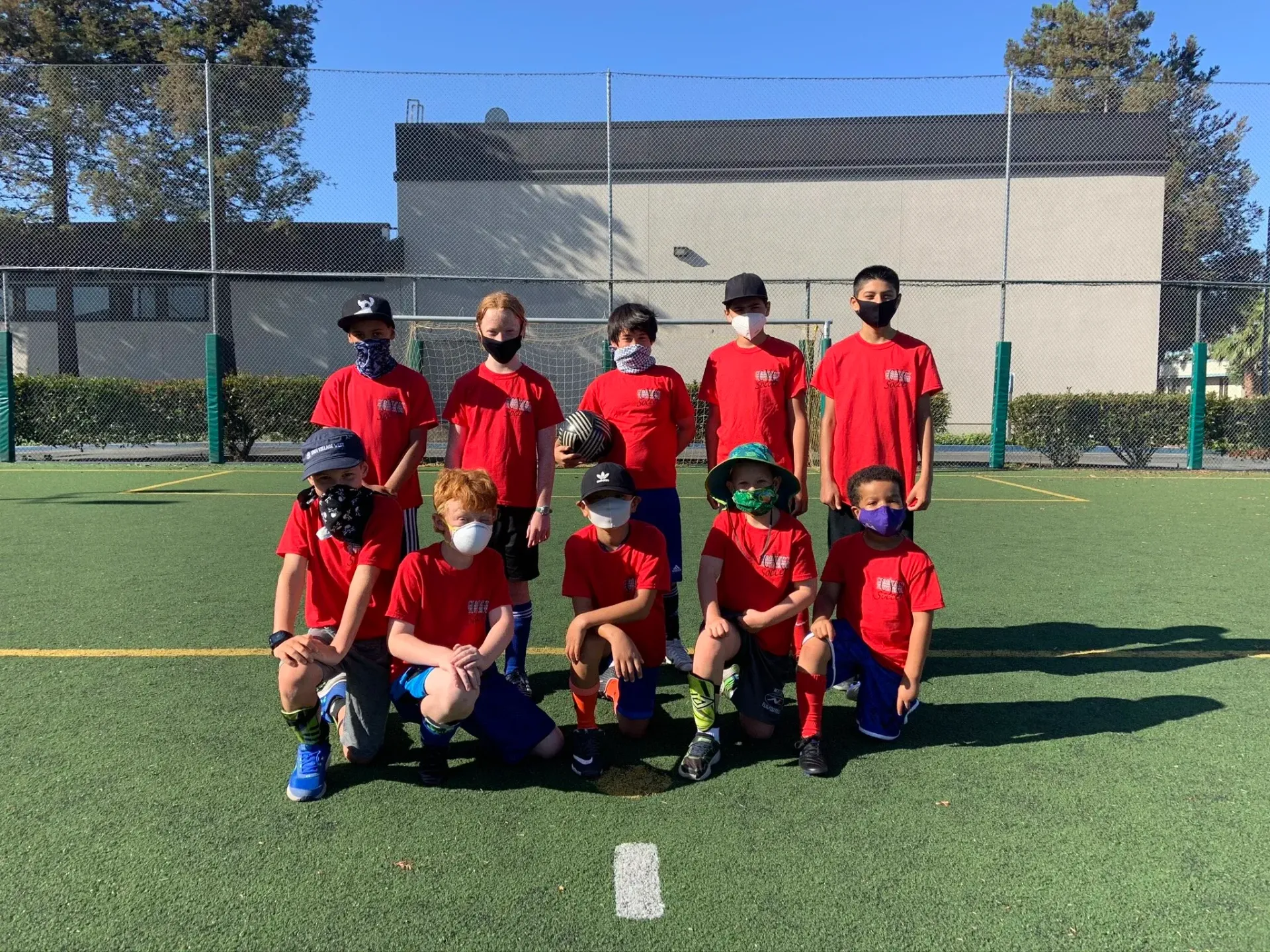 Soccer team poses on a green field wearing red shirts and masks. A ball is held by a player.