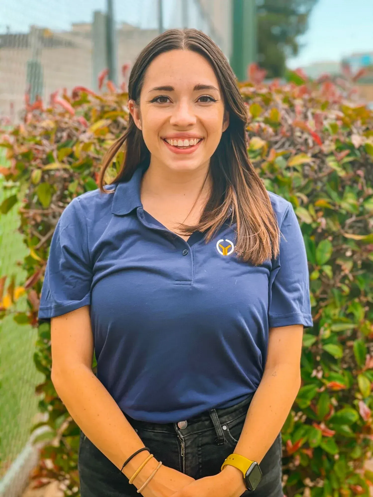 Woman in a blue polo shirt smiles, posing outdoors with green foliage in background.