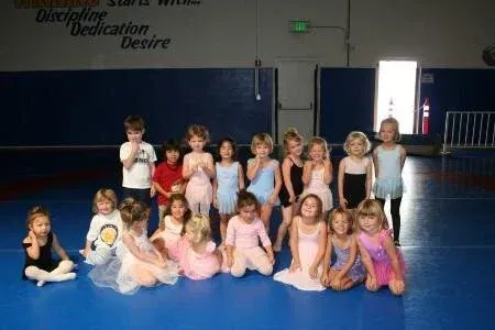 Children in ballet attire pose on a blue mat. Some are kneeling, smiling. A gym setting with motivational text on the wall.