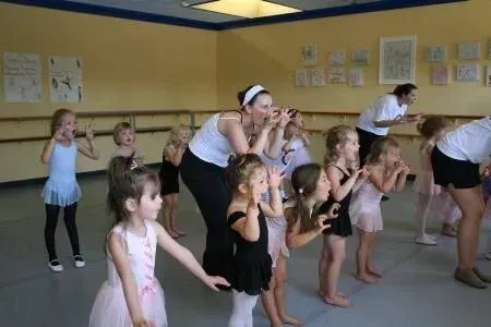 Children in leotards practice dance moves with instructors in a studio.