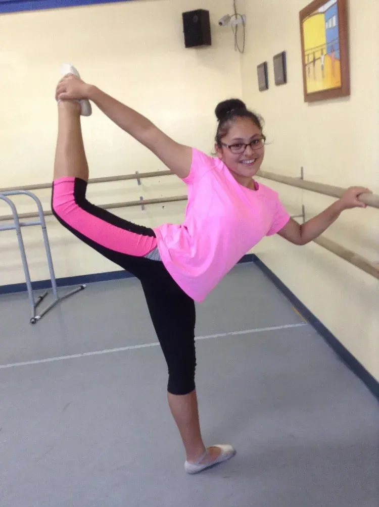 A person in pink top and black leggings does a ballet pose in a studio, holding one foot up with a smiling expression.