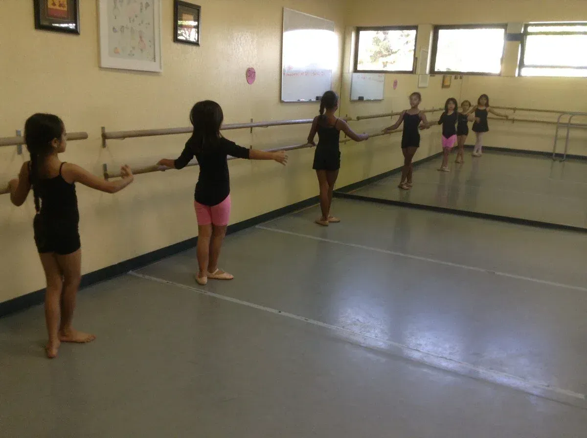 Young dancers practice ballet at a barre in a studio, arms outstretched.