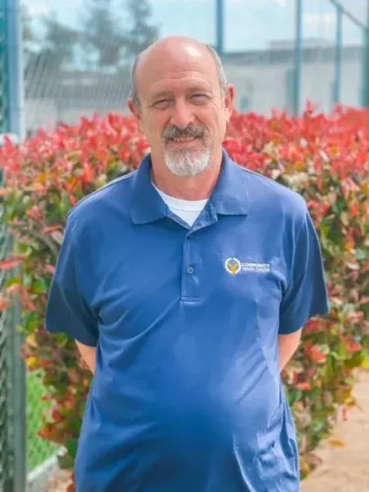 Man with gray beard, wearing a blue polo shirt, standing in front of red bushes.