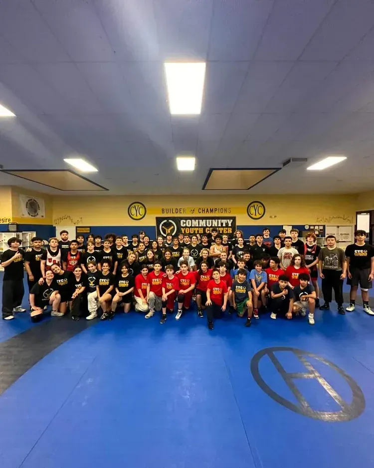 Group of people in a wrestling gym wearing various colored shirts on a blue mat.