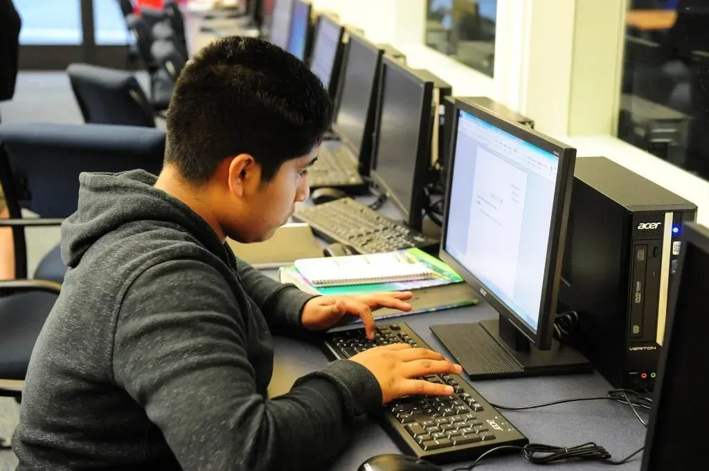 Person typing on a computer in a computer lab. Black keyboard and monitor, gray hoodie.