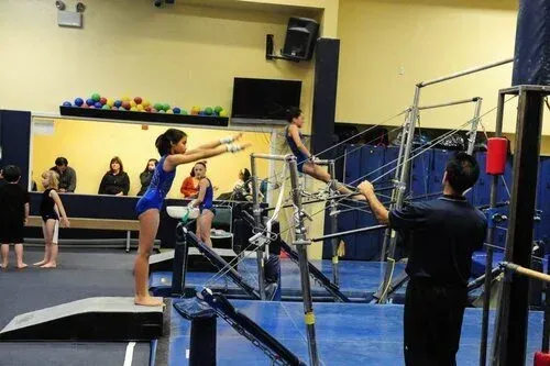 Gymnast practicing on uneven bars, another gymnast on bars, coach, and observers in gym.