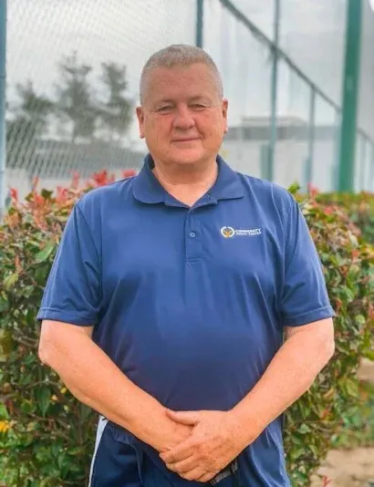 Man in a navy blue polo shirt stands outdoors with arms crossed. Green bushes and a fence are in the background.