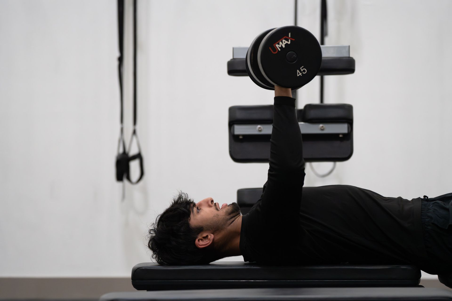 Person lying on a bench doing a dumbbell press in a gym, wearing black clothing.