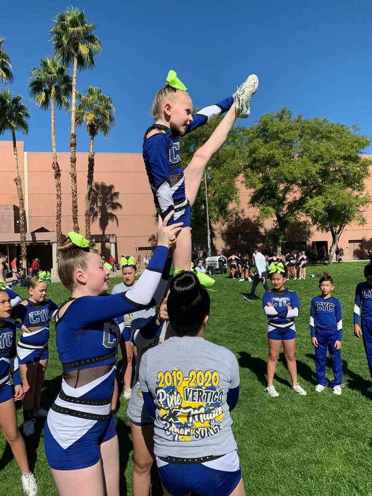 Cheerleaders in blue and white uniforms perform a stunt on a grassy field with palm trees.