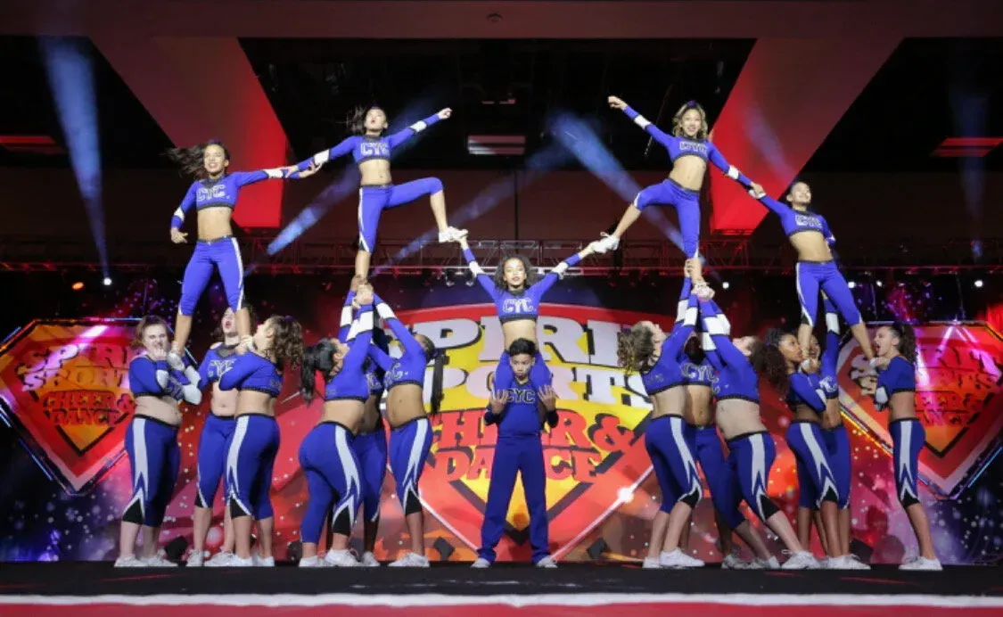 Cheerleading team in blue uniforms performing a pyramid stunt on a stage with a logo backdrop and spotlights.