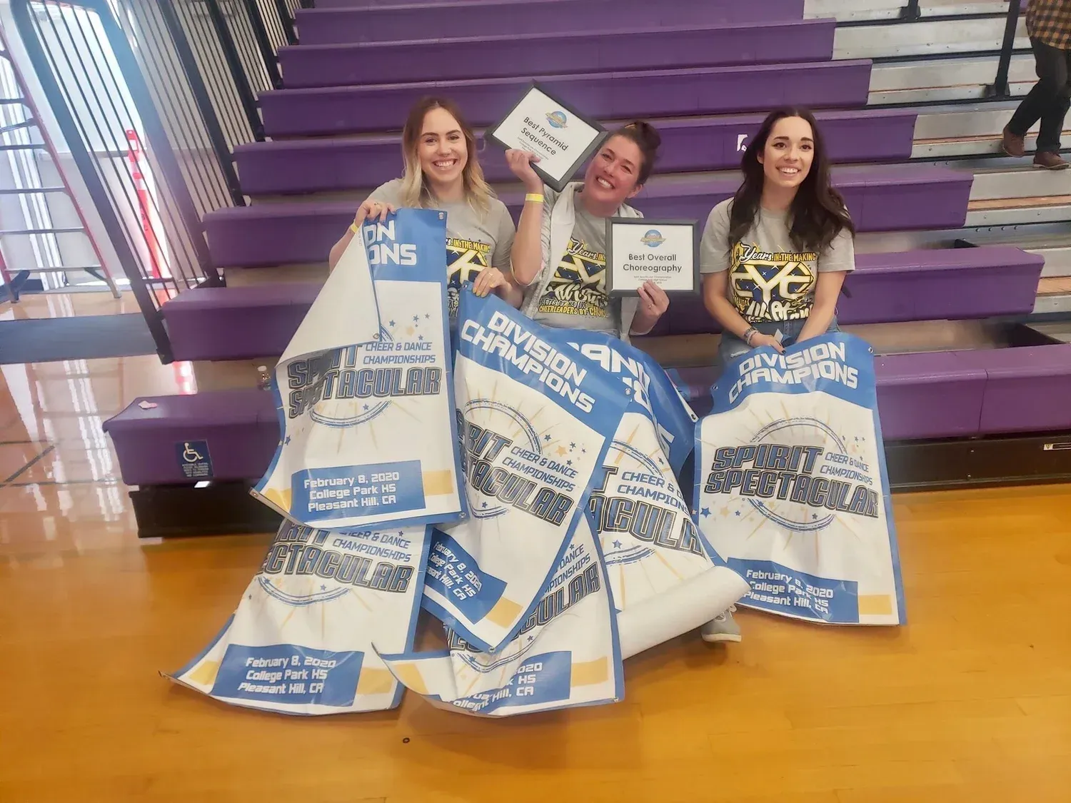 Three women holding champion banners and certificates, sitting on purple bleachers.