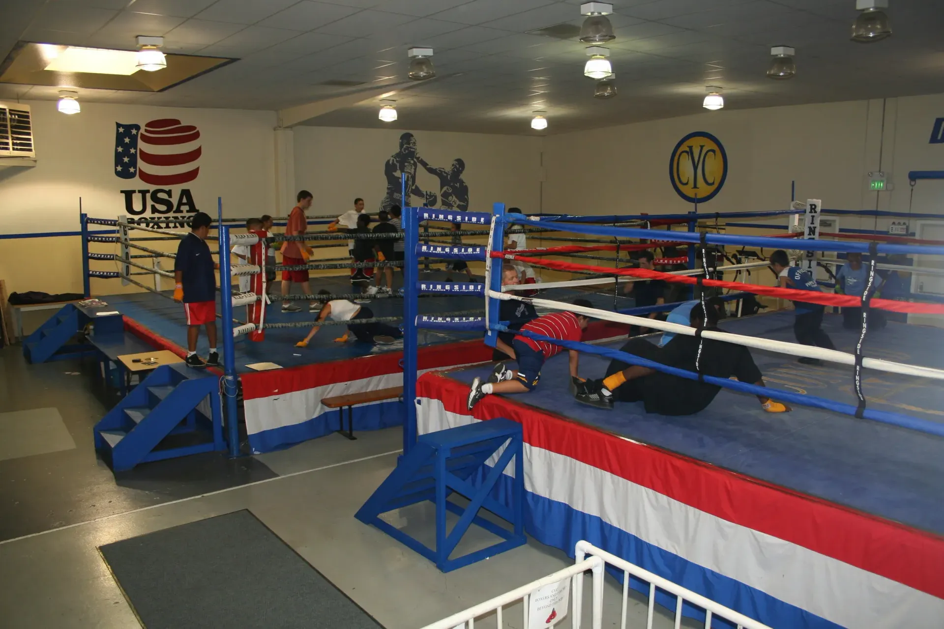 Boxing gym with multiple rings and people training. Rings have blue, red, and white trim; USA Boxing logo visible.