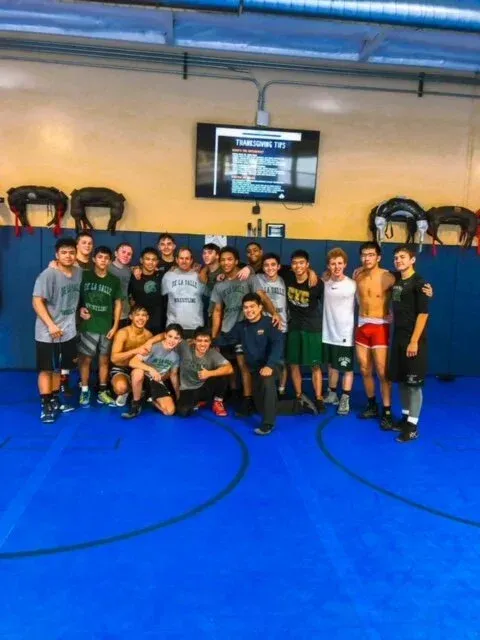Wrestling team poses on a blue mat in a gym. A TV is on the wall.
