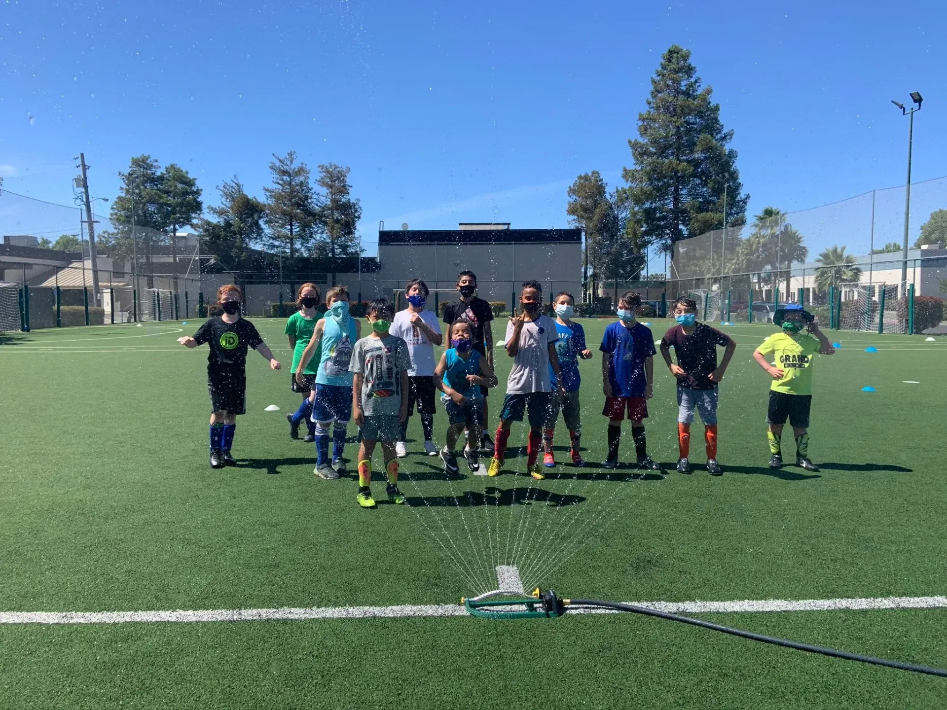Children on a soccer field, sprayed with water, many wearing masks, under a sunny sky.