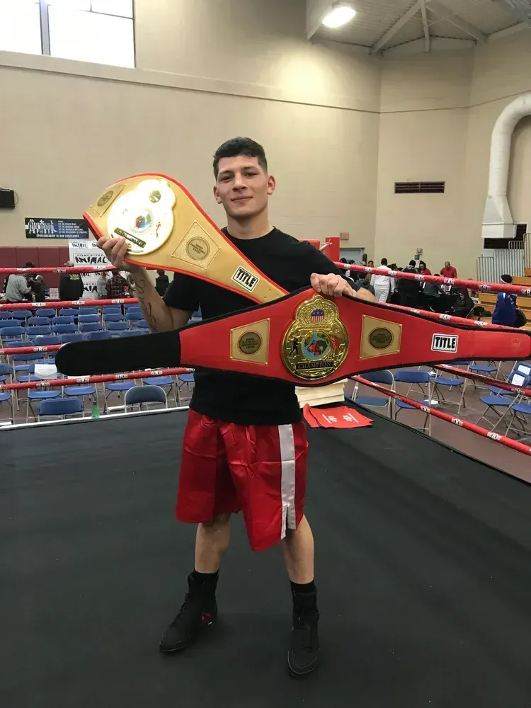 Boxer holding two championship belts in a boxing ring; wearing red shorts, black shirt, and shoes.