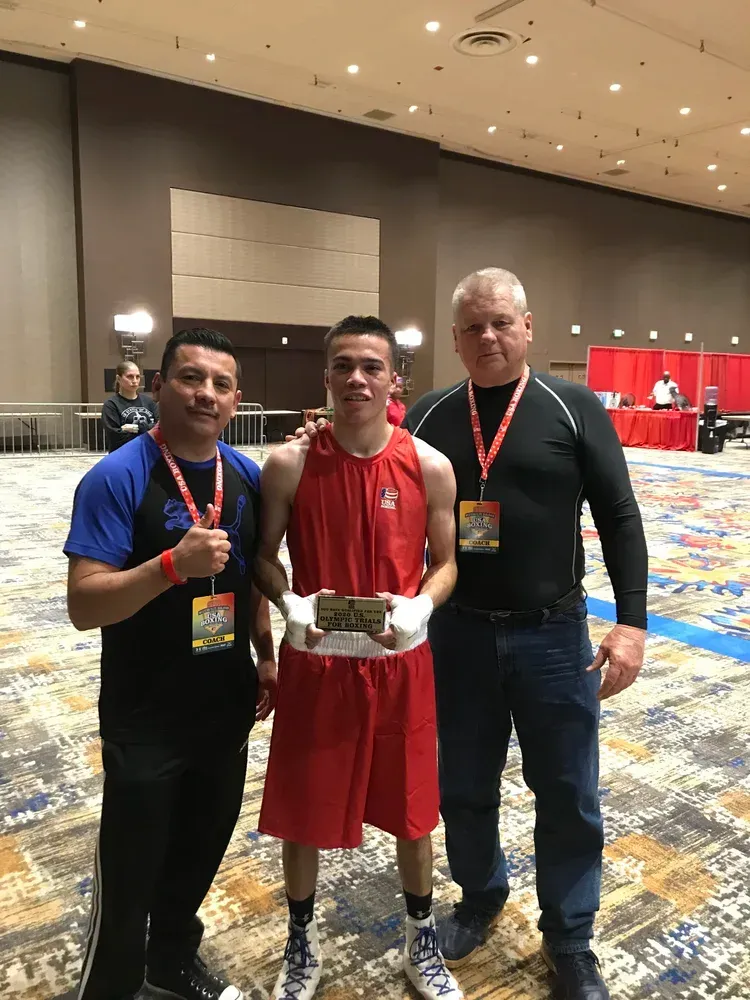 Boxer in red uniform holding a belt with two coaches, likely after a win at an event.