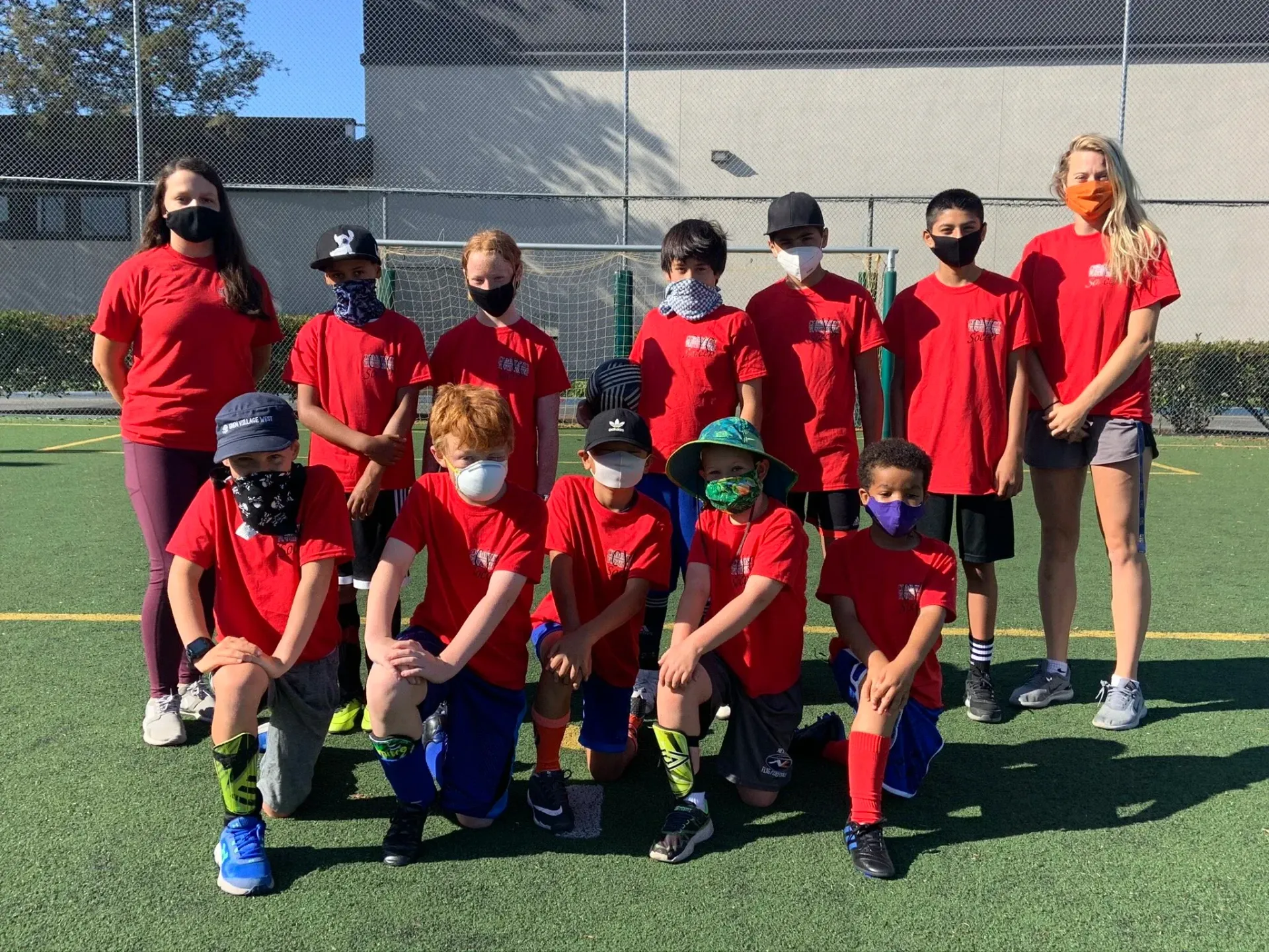 Soccer team in red shirts and masks on a green field. Two adults and ten children pose for the camera.