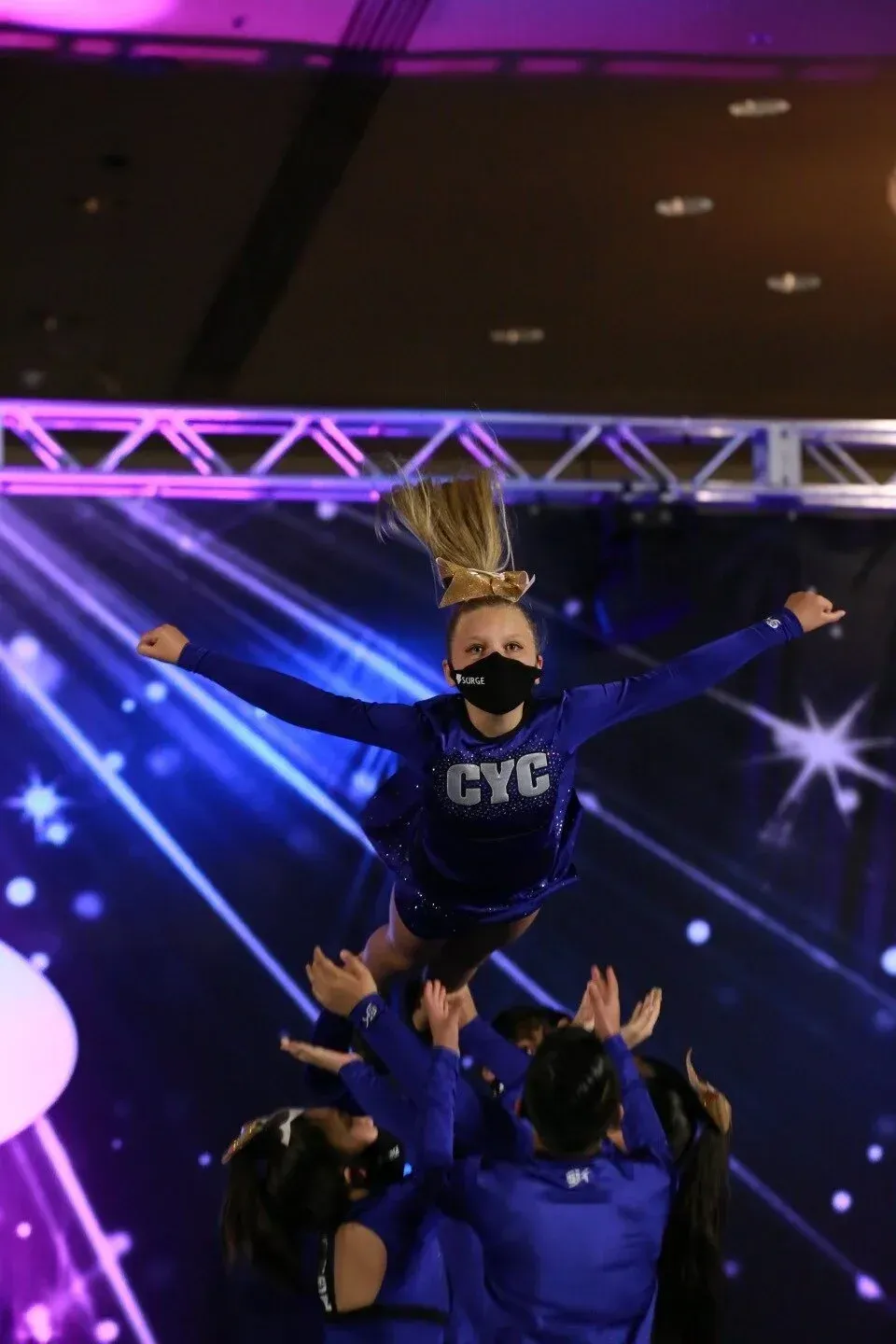 Cheerleader in a blue uniform is thrown in the air by her teammates, with a stage background and bright lighting.