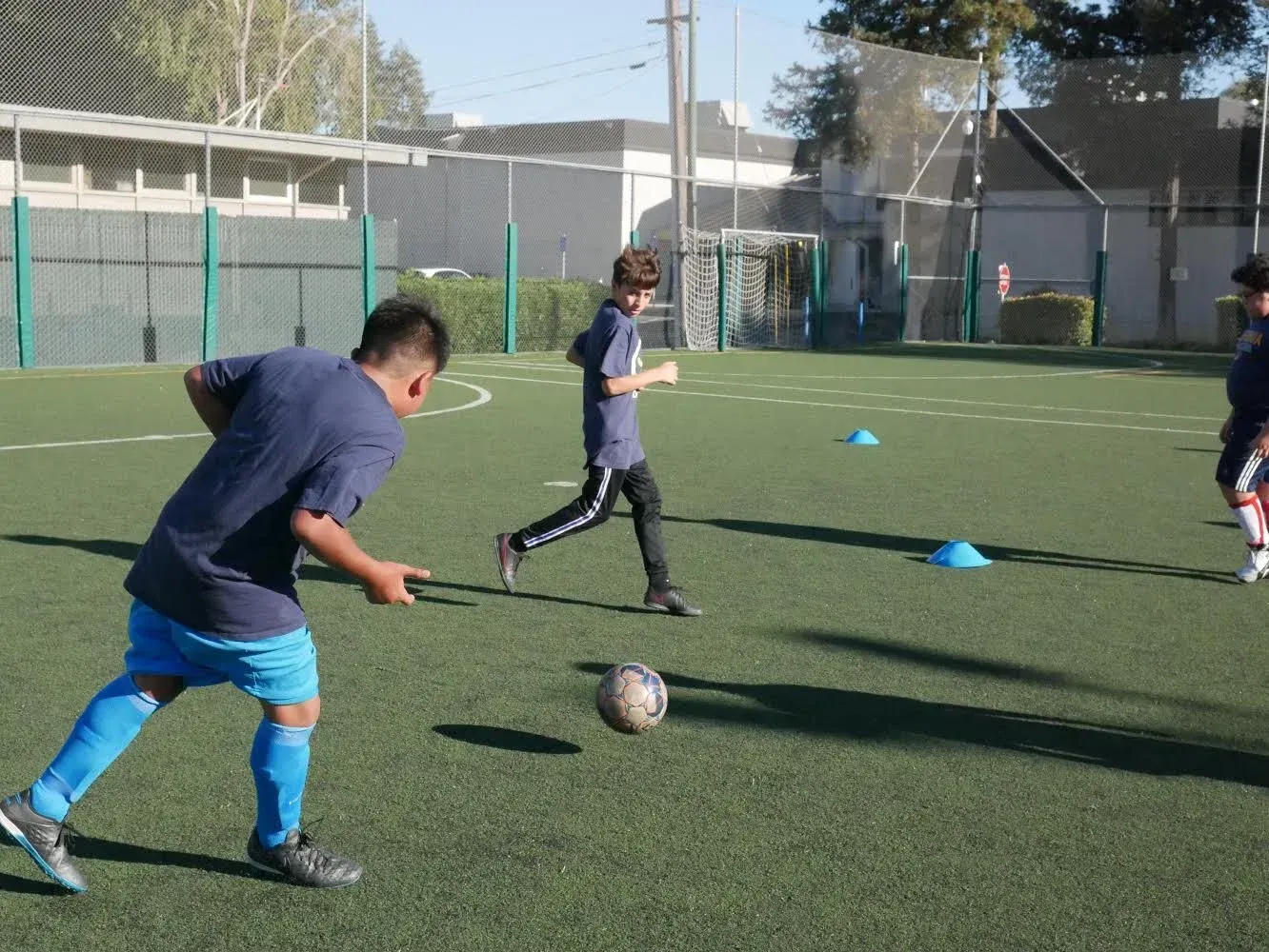 Players practice soccer on a green field. One kicks ball; another runs. Blue cones mark training area.