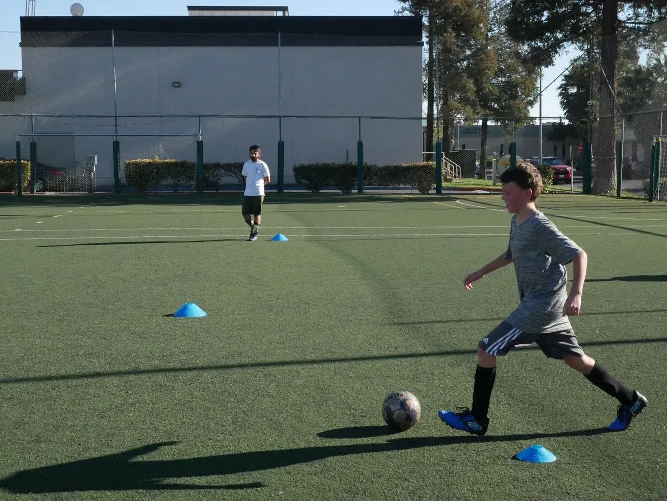 Boy kicking a soccer ball on a green field, two blue cones visible, another person in the background.