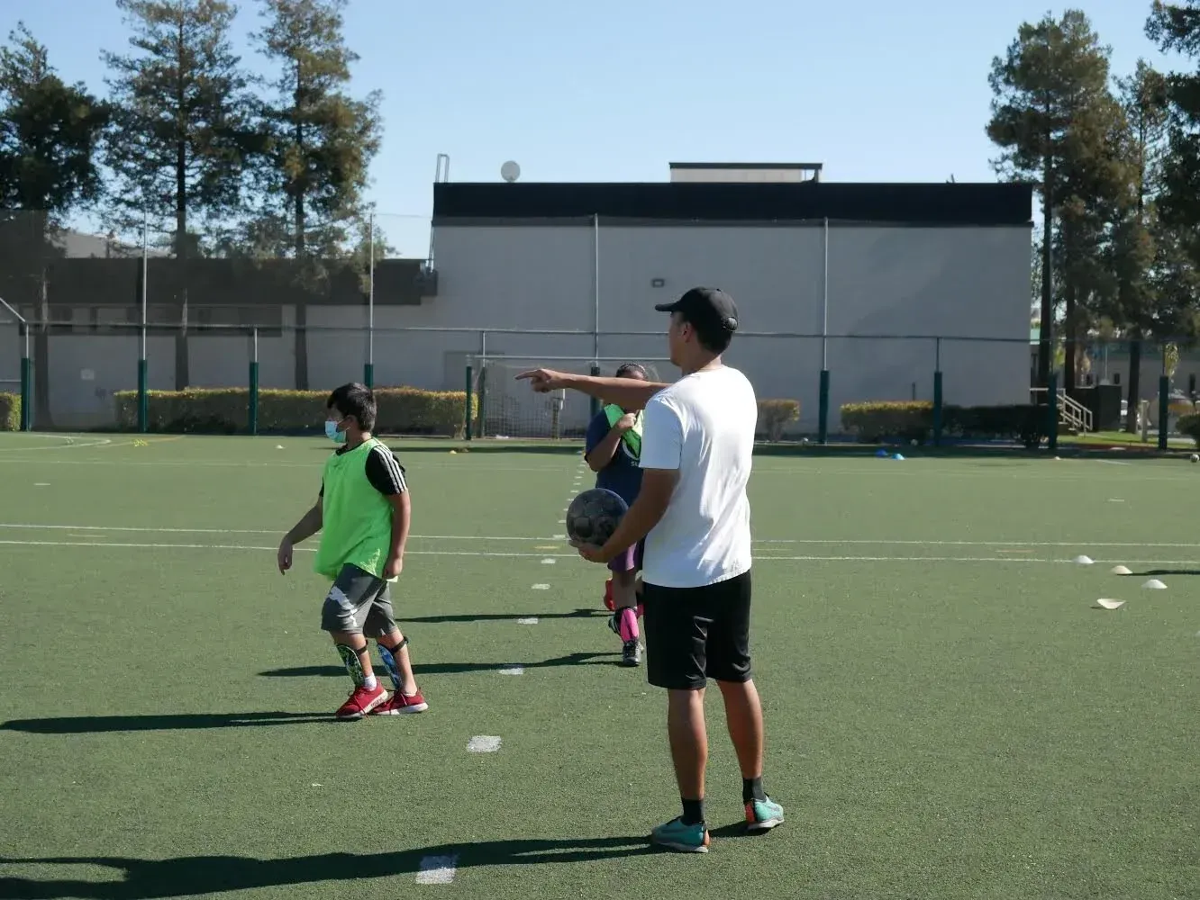 Man coaching children on a green field. He points, holding a ball, as kids in sports gear train.
