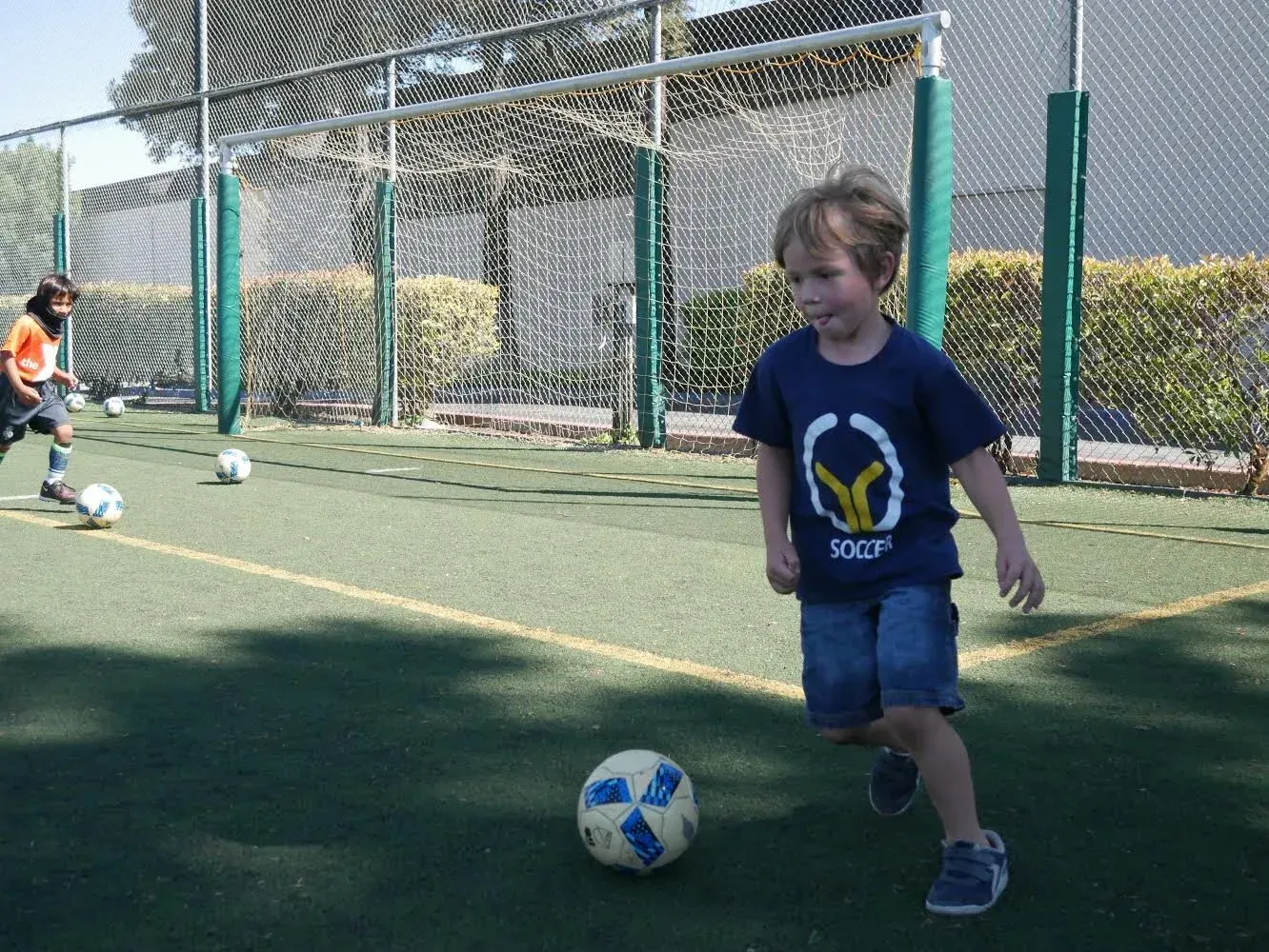 Boy in blue shirt dribbles soccer ball on green field; another player in background.