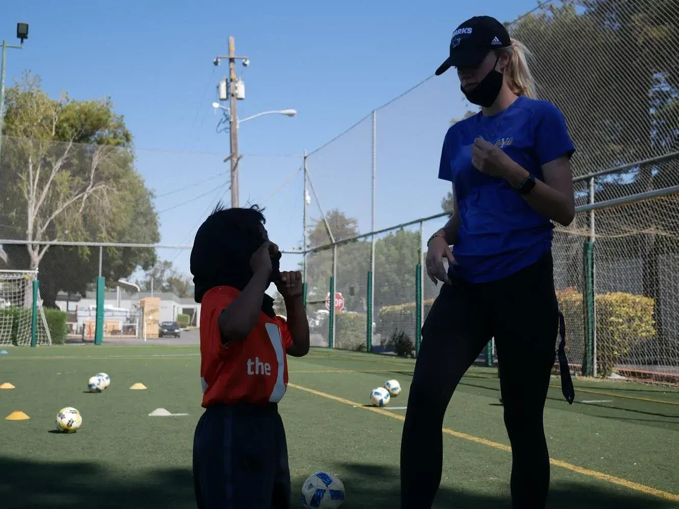 Child in soccer uniform talking to coach on a soccer field, several soccer balls, and cones are visible.