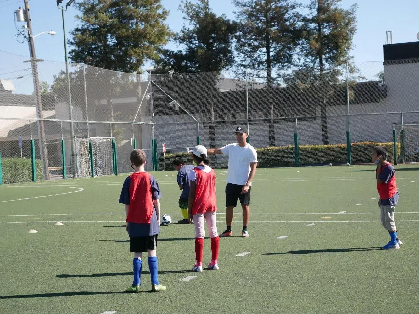 Soccer coach instructing children on a green field; players in red and blue practice gear.
