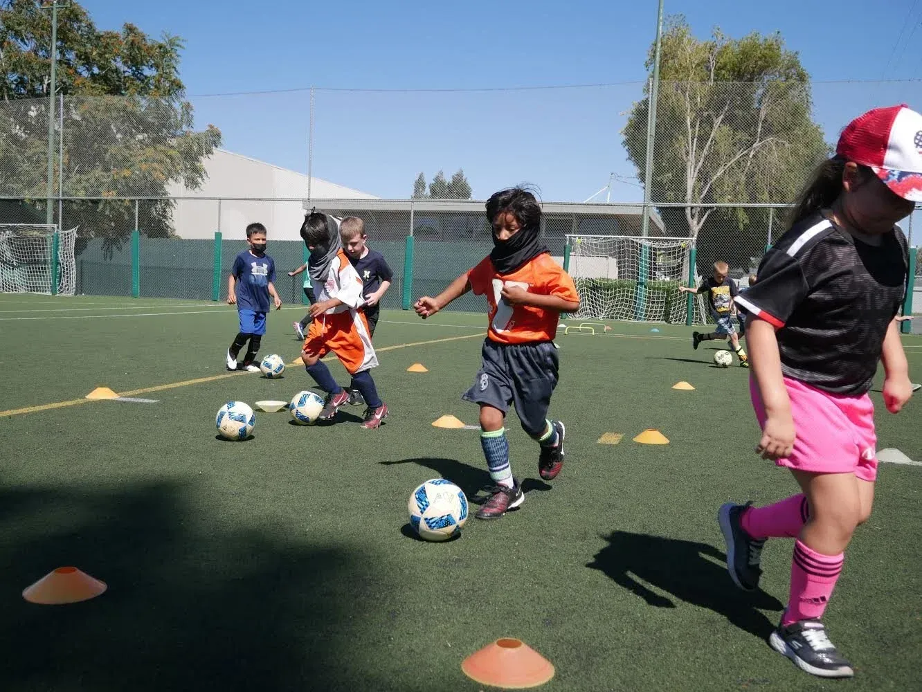 Children playing soccer on a green field with cones. One child in orange runs with the ball.