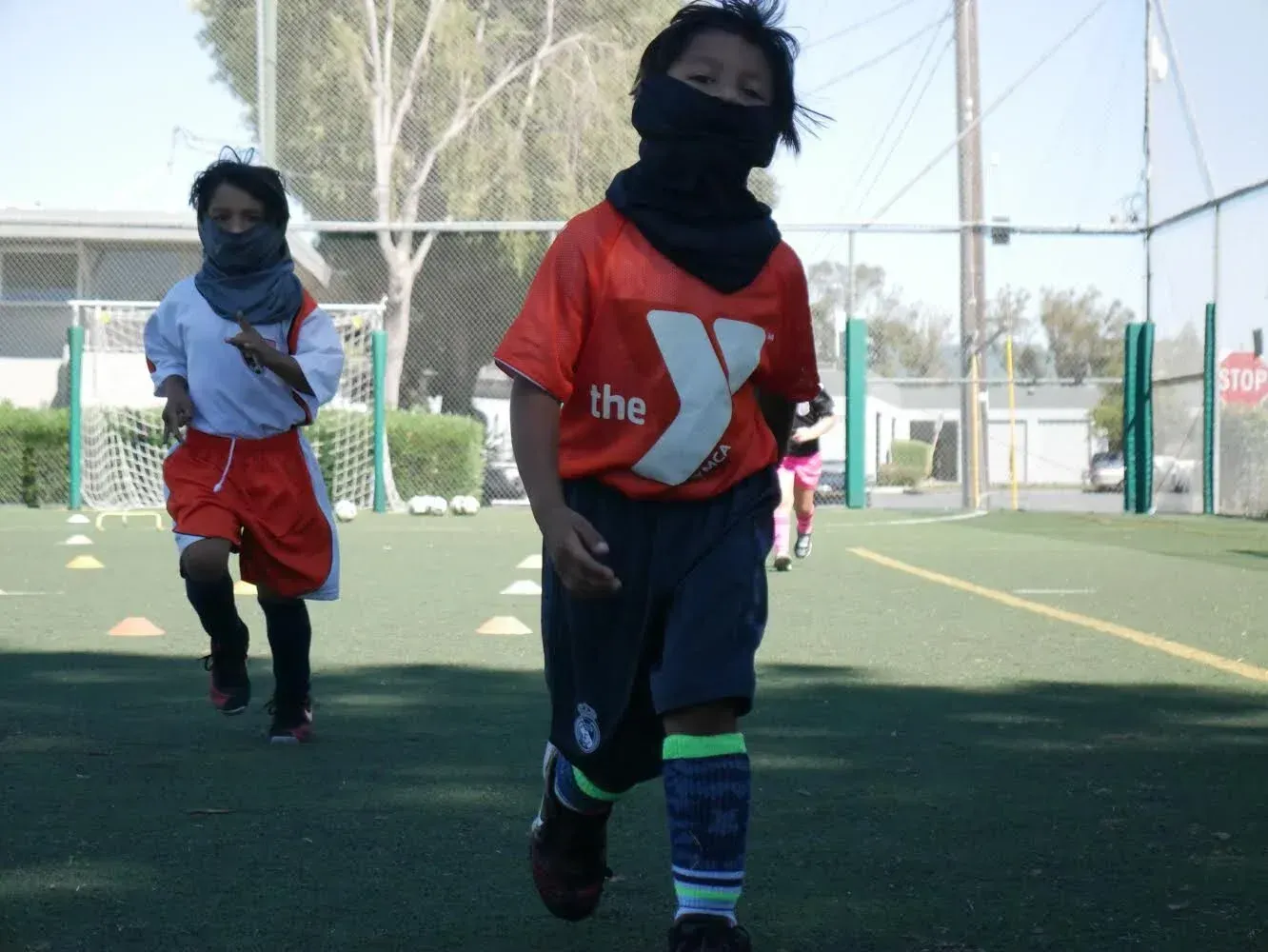 Two children running on a soccer field, faces covered by black scarves. One wears an orange shirt.