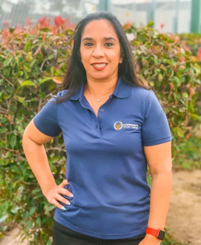 Woman in blue polo shirt stands with hand on hip outdoors, smiling.