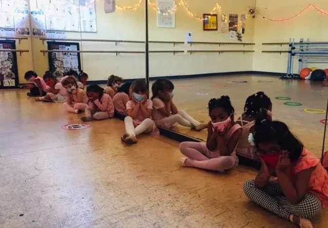 Children in pink dance outfits and masks sit on a dance studio floor.