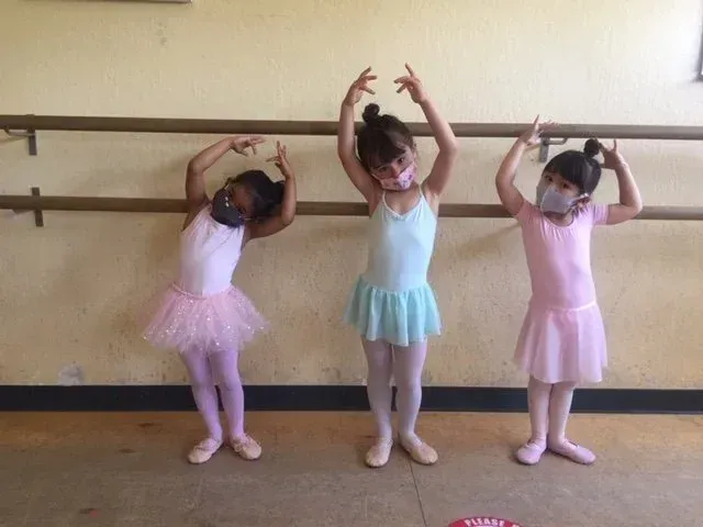 Three young children in ballet attire stand by a barre, arms raised, wearing masks.