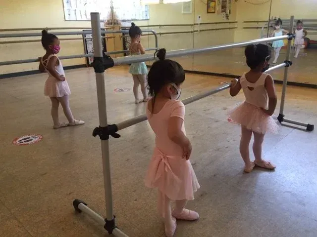 Young dancers wearing masks and pink tutus at a ballet barre in a studio.