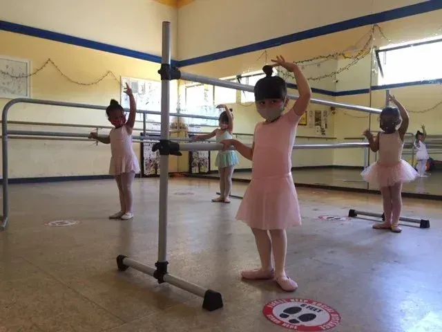 Children in pink ballet attire practice at the barre in a dance studio.