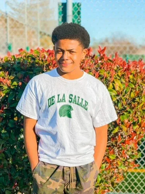 Teenager in a white t-shirt with green text, camo pants, smiling, outdoors.