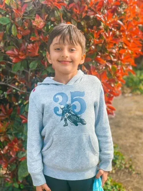 Boy in gray hoodie smiles in front of red-leafed bush.
