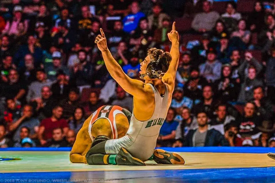 Wrestler in gray singlet celebrates victory, arms raised, opponent in orange singlet on mat, arena crowd in background.