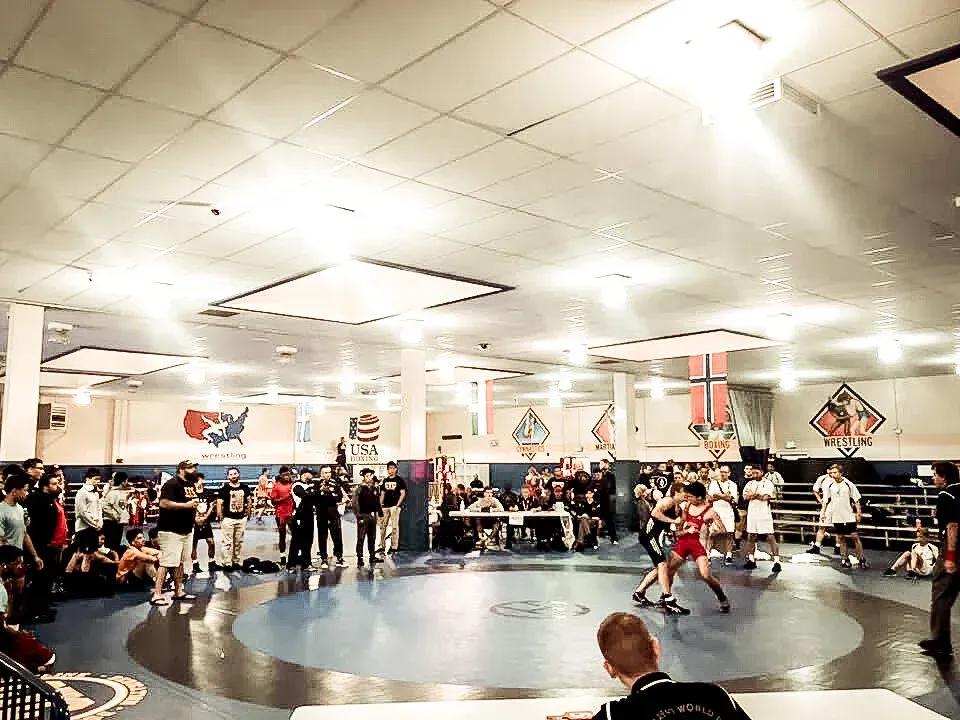 Wrestling match in progress on a blue mat, surrounded by spectators and officials in a brightly lit gym.