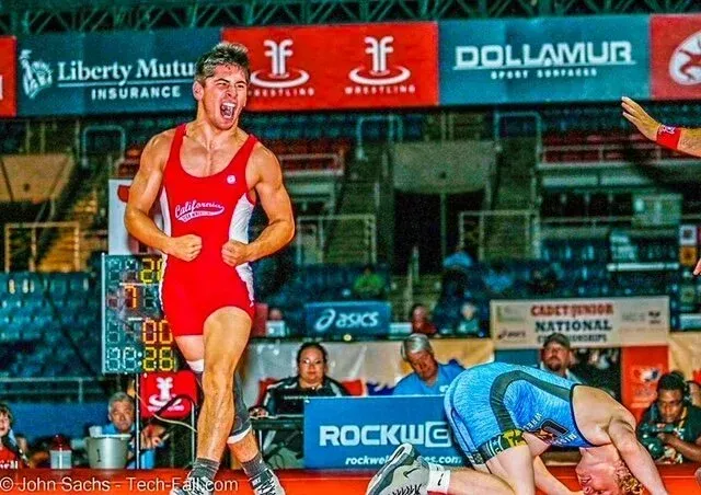 Wrestler in red singlet celebrating a victory as opponent lies on mat, crowd in background.