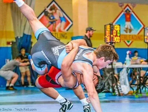 Wrestlers in action: One in a light blue singlet, executing a takedown on an opponent in a red singlet. Indoor setting.
