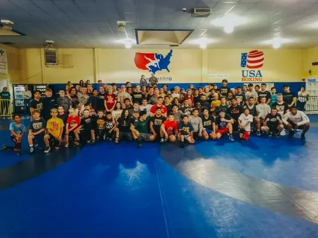Group of people, mostly young, on a blue wrestling mat in a gym. USA Wrestling logo on wall.