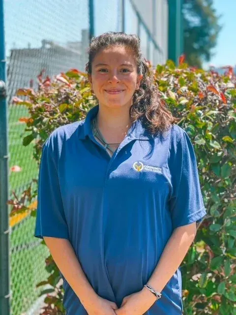 Woman wearing a blue polo shirt smiles outside near green bushes.